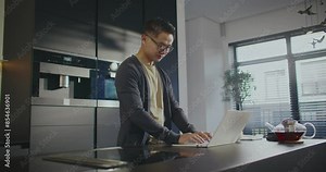 Handsome Asian man using laptop during his work. Working remotely with computer. Taking sip of hot drink. Feeling fresh during busy morning. Typing information on keyboard. Pressing keys.