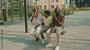 Happy African American family with cool preteen son chatting and eating ice-cream while sitting on playground swing outside their house on warm summer day