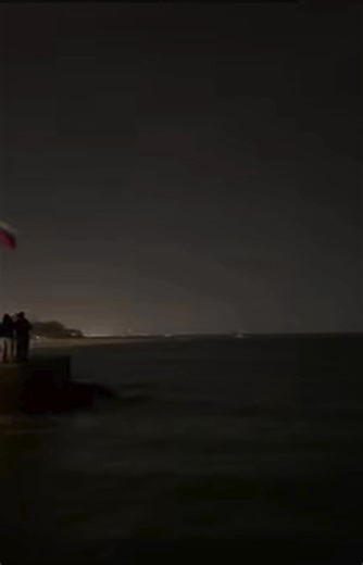 A tense nighttime phone recording from Venezuela captures civilians near the coast as a sudden military flyover turns a quiet shoreline into chaos. The video opens on the dark sea, waves barely audible, when the distant thunder of jet engines rushes in unexpectedly. Fighter jets then rip along the coastline, their flashing lights cutting through low clouds and reflecting across the water, sparking loud shouts and confusion. After the jets disappear, a brief silence settles in—only to be shattere