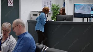 Patient talking to receptionist at hospital reception desk, asking for help with medical appointment and treatment. African american woman chatting with clinical staff in waiting area lobby.