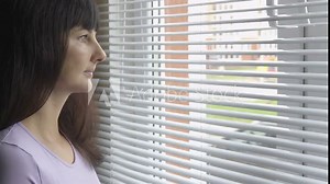 Young beautiful curious woman looks through the window through blinds. The girl is watching out the window. The brunette woman.